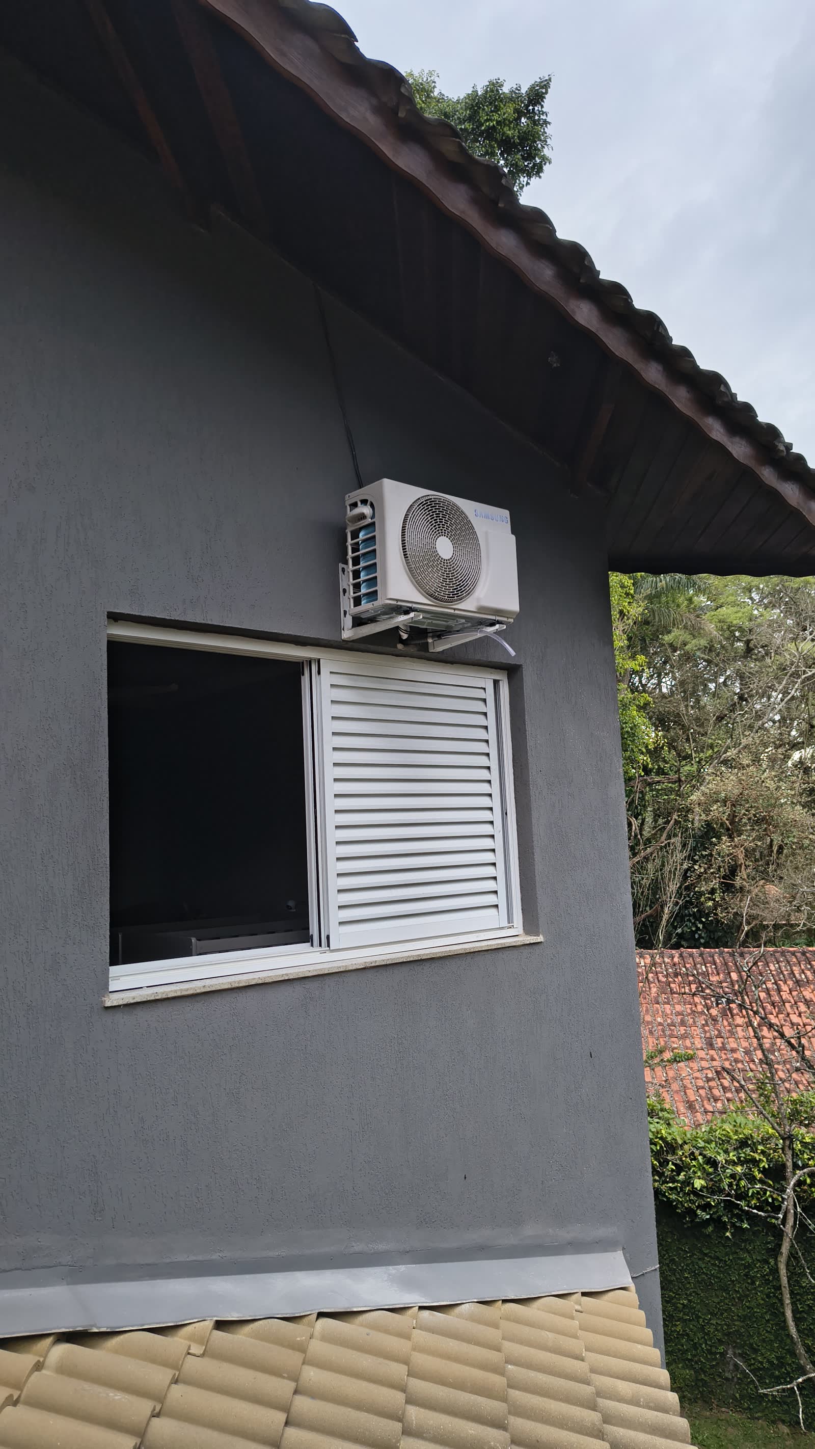 Condensadora fixada na parede externa de uma casa cinza, acima da janela